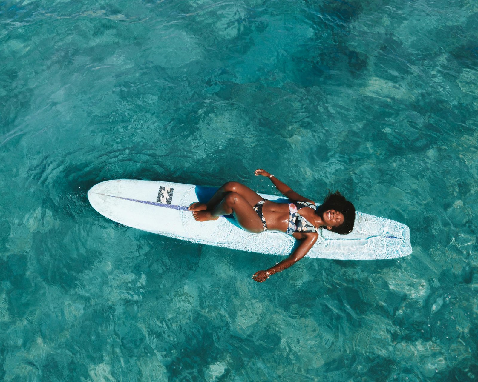 A woman in a bikini lying on a surfboard in clear blue water, enjoying a sunny day.
